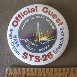 May include: White round button with red and blue text that reads "Official Guest NASA Ames-Dryden STS-26 Edwards AFB". The button features a graphic of a rocket launch with a yellow sun and red and white clouds.