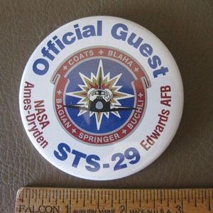 May include: White round button with blue and red text that reads "Official Guest NASA Ames-Dryden Edwards AFB STS-29". The button features a blue and white circular graphic with a gold starburst and a space shuttle.
