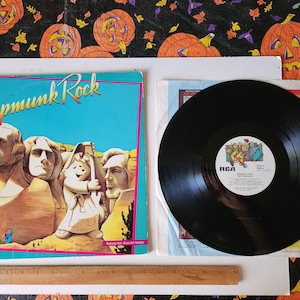May include: A vintage vinyl record album cover with a blue background and the title "Chipmunk Rock" in white text. The album cover features a cartoon image of four chipmunks dressed as presidents, standing in front of a mountain range. The album is accompanied by a black vinyl record with a white label that reads "Chipmunk Rock - The Chipmunks".