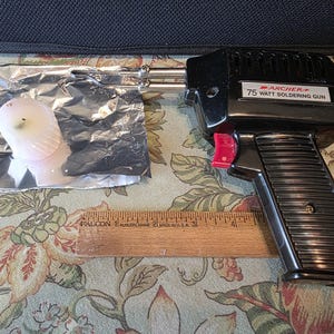 May include: A black Archer 75-watt soldering gun with a red trigger, next to a wooden ruler and a small object wrapped in foil. The soldering gun has a ribbed handle and the text "75 Watt Soldering Gun".