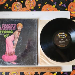 May include: A vinyl record album cover featuring a woman in a pink dress and the text "Rusty Warren Bottoms Up" with the words "For Adults Only" in smaller font.