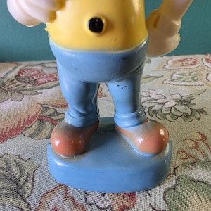 May include: A yellow and blue plastic figurine of a cartoon character wearing a yellow shirt with black buttons, blue pants, and brown shoes. The figurine is standing on a blue base.