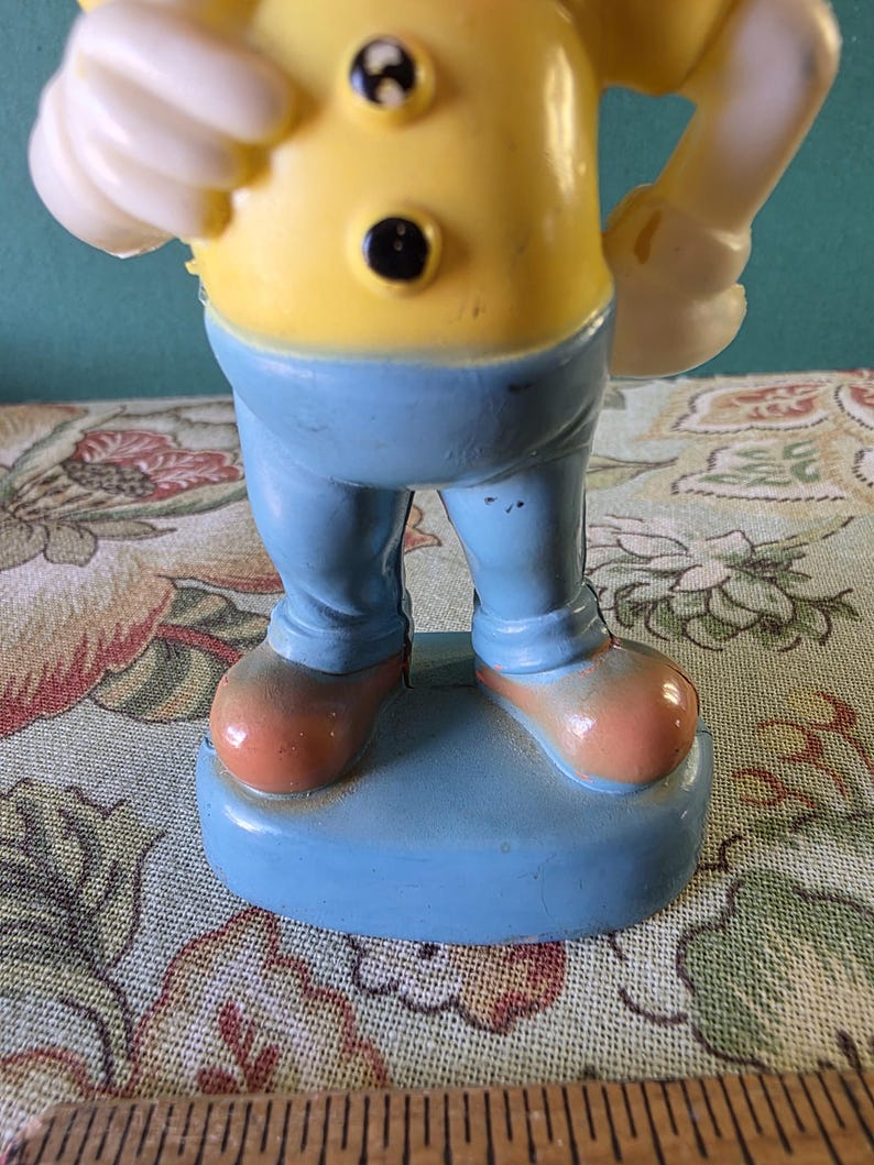 May include: A yellow and blue plastic figurine of a cartoon character wearing a yellow shirt with black buttons, blue pants, and brown shoes. The figurine is standing on a blue base.