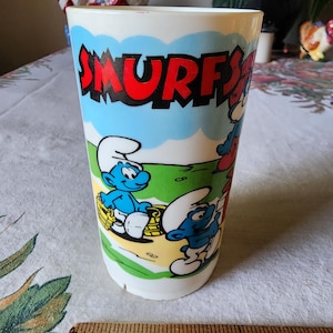 May include: A white plastic Smurfs drinking cup with a blue Smurf and a red Smurf on a green background. The cup has the word "SMURFS" in red letters on the front.