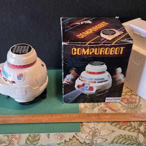 May include: A vintage Compurobot toy, white with red and black accents, sits next to its original box. The box features the Compurobot logo and an image of the robot. A ruler and instruction manual are also present.