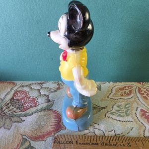 May include: A vintage plastic Mickey Mouse figurine wearing a yellow shirt, blue pants, and a red bow tie. The figurine is standing on a blue base.