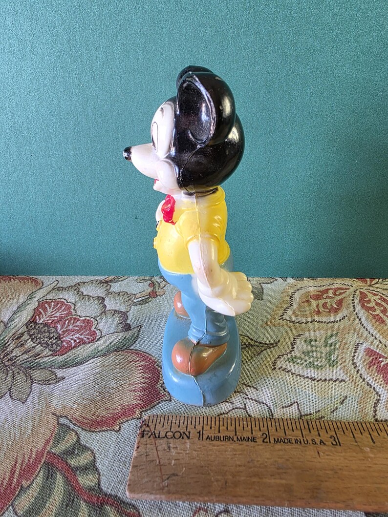 May include: A vintage plastic Mickey Mouse figurine wearing a yellow shirt, blue pants, and a red bow tie. The figurine is standing on a blue base.