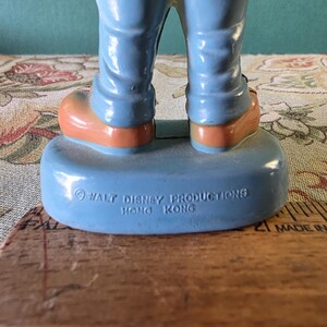 May include: A blue plastic figurine of a cartoon character wearing blue pants and brown shoes. The figurine is standing on a blue base with the text "&copy; WALT DISNEY PRODUCTIONS HONG KONG" printed on it.