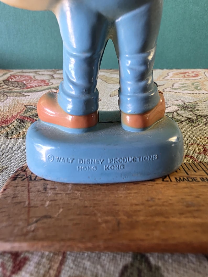 May include: A blue plastic figurine of a cartoon character wearing blue pants and brown shoes. The figurine is standing on a blue base with the text "&copy; WALT DISNEY PRODUCTIONS HONG KONG" printed on it.