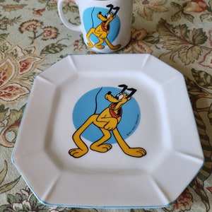 May include: A white ceramic plate with a blue rim and a cartoon image of Pluto the dog. The plate is square with rounded corners. The image of Pluto is on a blue circle with the text "© WALT DISNEY PRODUCTIONS". A white ceramic mug with a blue rim and a cartoon image of Pluto the dog is also included.