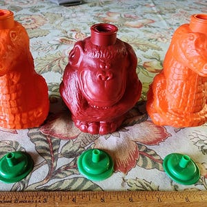 Puede incluir: Tres botellas de plástico exprimibles con forma de animal con tapas verdes. Las botellas son naranja, roja y naranja. Las botellas están sobre un mantel floral.