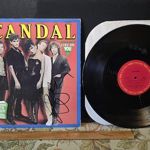 May include: A vintage vinyl record album by the band Scandal. The album cover features a group photo of the band members with the word "SCANDAL" in large yellow letters. The album is next to a black vinyl record.
