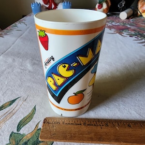 May include: A white plastic cup with a Pac-Man design. The cup has a blue, yellow, red, and orange design with the words "Amazing" and "Pac-Man" on it. The cup is 17 cm tall.
