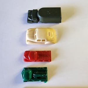 May include: Four toy vehicles, a green truck, a white car, a red truck, and a green truck, are lined up vertically on a white surface. A wooden ruler with the words "FALCON" and "AUBURN, MAINE" is visible at the bottom of the image.