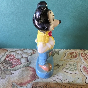 May include: A vintage plastic figurine of Mickey Mouse wearing a yellow shirt, blue jeans, and a red bow tie. The figurine is standing on a blue base.