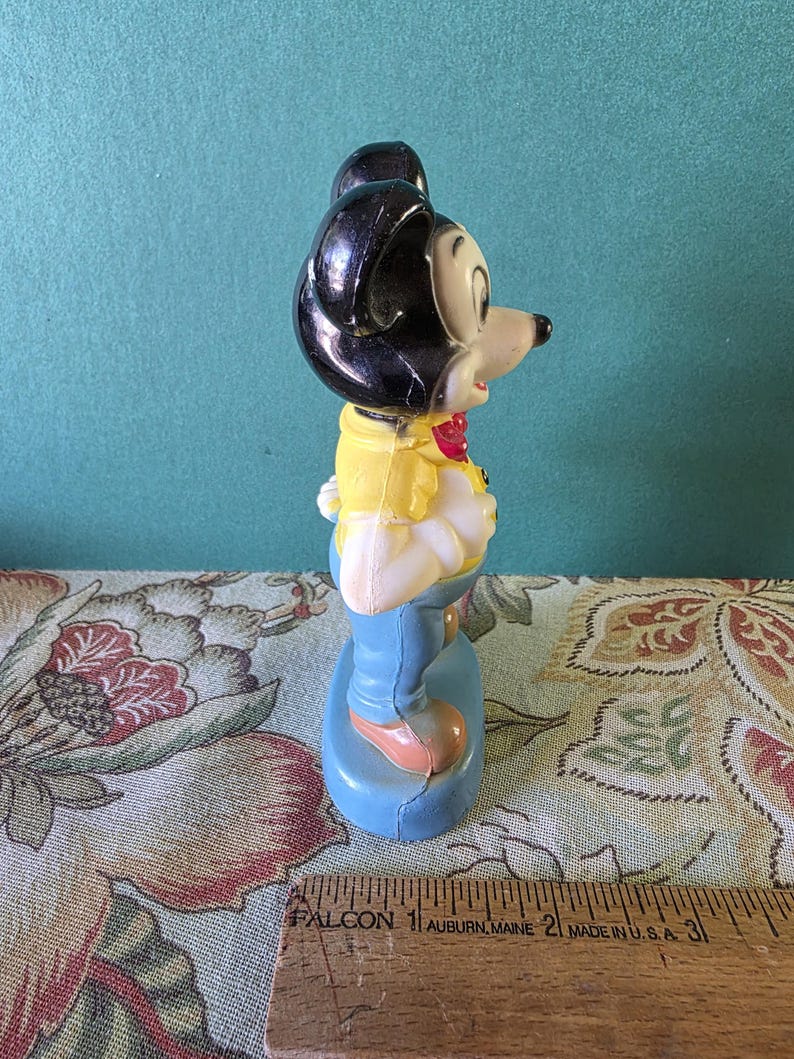May include: A vintage plastic figurine of Mickey Mouse wearing a yellow shirt, blue jeans, and a red bow tie. The figurine is standing on a blue base.