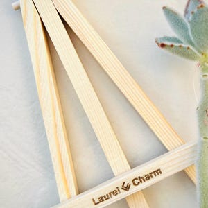 Könnte beinhalten: Ein kleiner, unfertiger Holzstaffelei mit dem Text "Laurel Charm" auf der Vorderseite eingraviert. Die Staffelei ist aus hellfarbenem Holz gefertigt und hat ein dreibeiniges Design. Im Hintergrund ist eine Sukkulentenpflanze zu sehen.