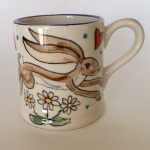 Może przedstawiać: Biały kubek ceramiczny z ręcznie malowanym wzorem przedstawiającym brązowego królika biegnącego przez pole białych stokrotek z czerwonymi sercami w tle.