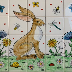 May include: A hand-painted ceramic tile depicting a brown rabbit sitting in a field of green grass with yellow flowers, blue flowers, and a snail. The tile is decorated with a dragonfly, a bee, and butterflies.