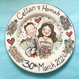 May include: A white ceramic plate with a blue rim, featuring a hand-painted illustration of a bride and groom with a dog. The plate is decorated with flowers, hearts, and the text "Cellan & Hannah 30th March 2024".