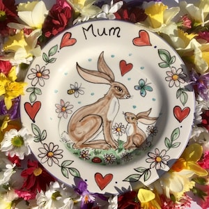 May include: A white ceramic plate with a hand-painted design of two hares in a field of flowers. The plate has a blue rim and the word "Mum" written in black lettering. The plate is surrounded by a variety of flowers.