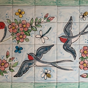 May include: A hand-painted tile mural featuring three blue birds with red breasts flying amongst pink flowers and green leaves. The tile is decorated with a dragonfly, bees, and butterflies. The tile is signed "Kate Glanville".