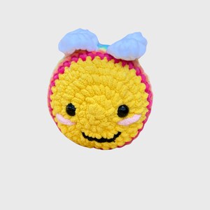 May include: A handmade, crocheted yellow smiley face with pink and white accents. The smiley face has black eyes, pink cheeks, and a curved black mouth. Two white cloud-like shapes are attached to the top of the smiley face.