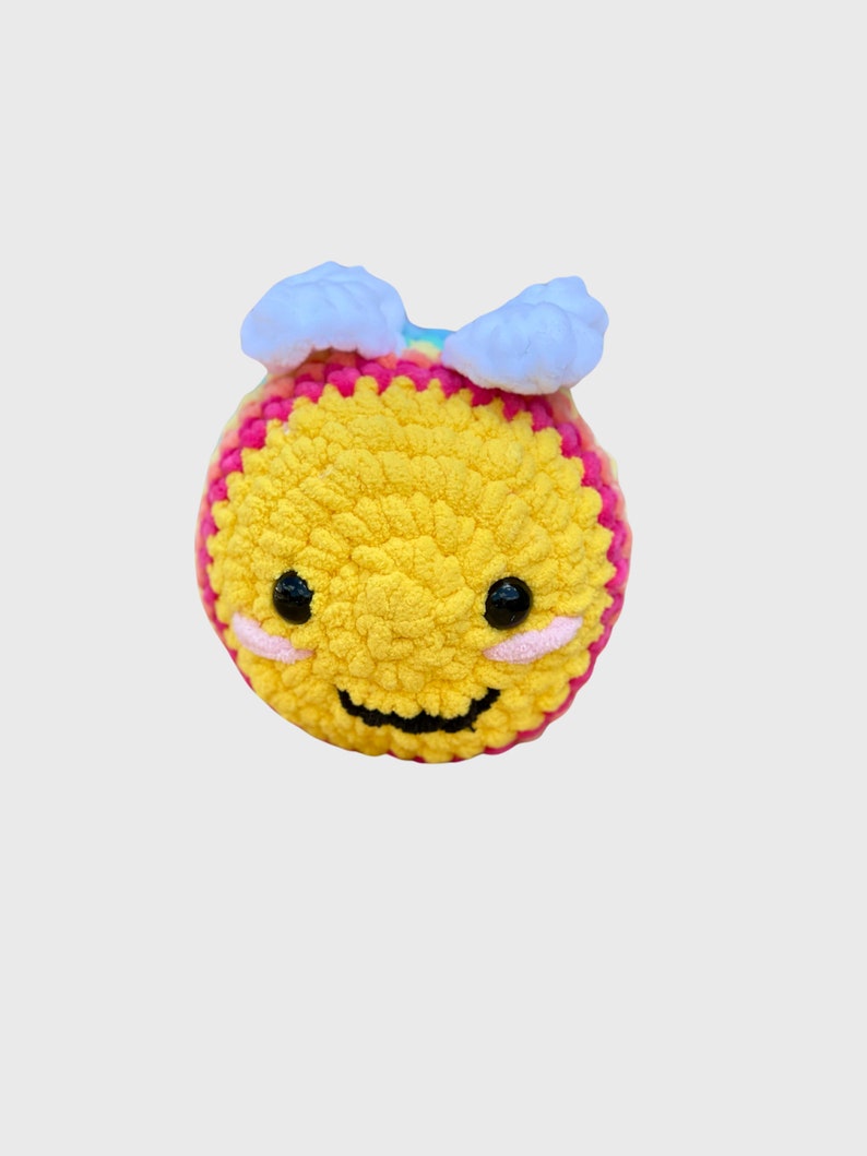 May include: A handmade, crocheted yellow smiley face with pink and white accents. The smiley face has black eyes, pink cheeks, and a curved black mouth. Two white cloud-like shapes are attached to the top of the smiley face.