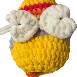 May include: A handmade, yellow crocheted bee with white wings and a rainbow-colored stripe across the top. The bee has an orange stitch marker attached to a yellow yarn tail. The bee is made of soft, plush yarn.