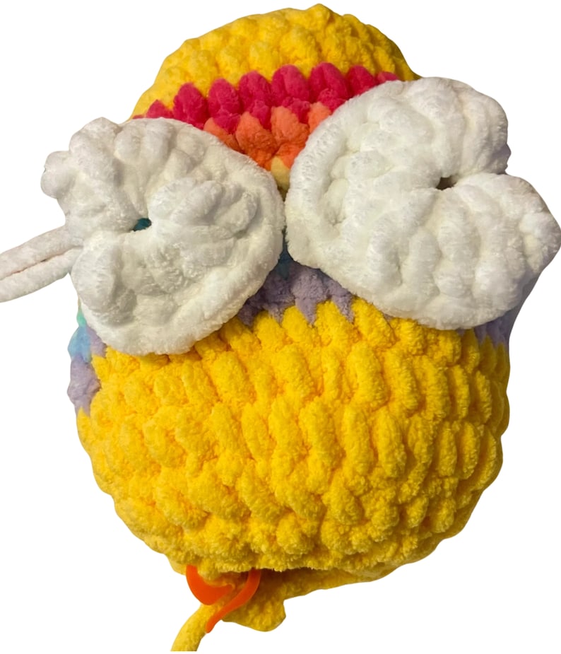 May include: A handmade, yellow crocheted bee with white wings and a rainbow-colored stripe across the top. The bee has an orange stitch marker attached to a yellow yarn tail. The bee is made of soft, plush yarn.