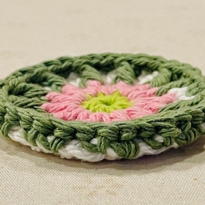 Pu&ograve; includere: Un sottobicchiere rotondo lavorato all'uncinetto con un motivo floreale. Il centro &egrave; di un verde lime brillante, circondato da strati di petali rosa tenue, un anello bianco e un bordo esterno in filato verde oliva testurizzato.