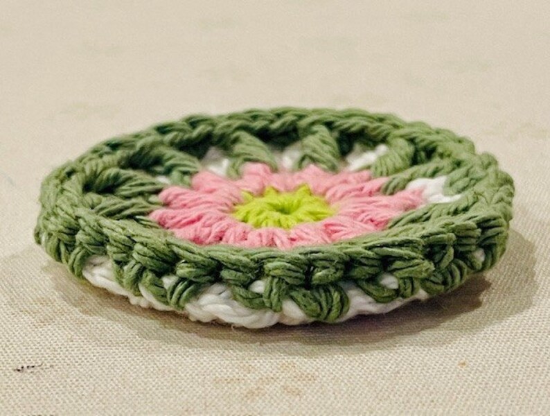 Pu&ograve; includere: Un sottobicchiere rotondo lavorato all'uncinetto con un motivo floreale. Il centro &egrave; di un verde lime brillante, circondato da strati di petali rosa tenue, un anello bianco e un bordo esterno in filato verde oliva testurizzato.
