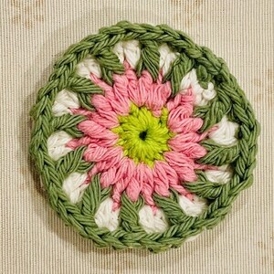 Pu&ograve; includere: Un sottobicchiere circolare all'uncinetto caratterizzato da un centro verde lime vibrante, circondato da strati di petali in filato rosa tenue, accenti bianchi e un bordo spesso e strutturato color verde oliva con punti visibili.