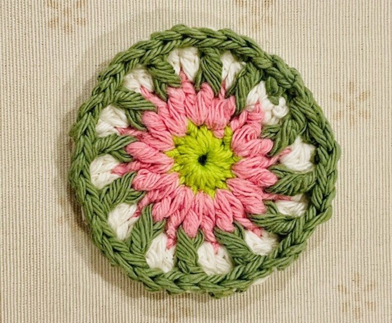 Pu&ograve; includere: Un sottobicchiere circolare all'uncinetto caratterizzato da un centro verde lime vibrante, circondato da strati di petali in filato rosa tenue, accenti bianchi e un bordo spesso e strutturato color verde oliva con punti visibili.