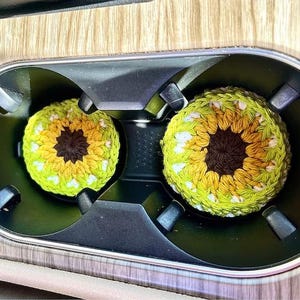 Pu&ograve; includere: Due sottobicchieri rotondi all'uncinetto con un design ispirato ai girasoli. Ogni sottobicchiere presenta un centro marrone scuro circondato da strati di petali in filato giallo e verde chiaro, creando un motivo floreale circolare per portabicchieri.