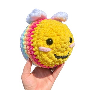 May include: A handmade, plush bee toy with a yellow body, rainbow stripes, and white cloud accents. The bee has black eyes, pink cheeks, and a stitched black smile. The toy is held in a hand against a white background.