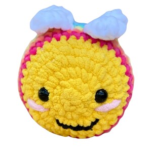 May include: A handmade, yellow crocheted bee with a smiling face, black eyes, and pink cheeks. The bee has white cloud-shaped accents on its head and a rainbow-colored stripe. The bee is a soft, plush toy.