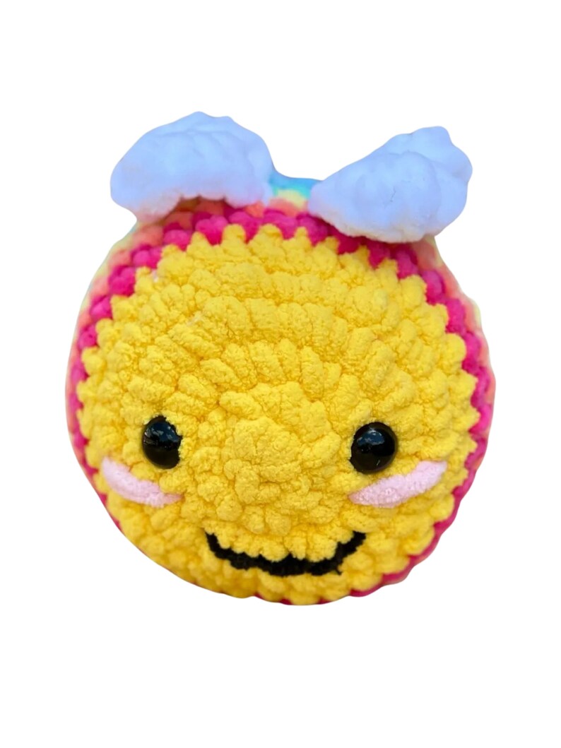 May include: A handmade, yellow crocheted bee with a smiling face, black eyes, and pink cheeks. The bee has white cloud-shaped accents on its head and a rainbow-colored stripe. The bee is a soft, plush toy.