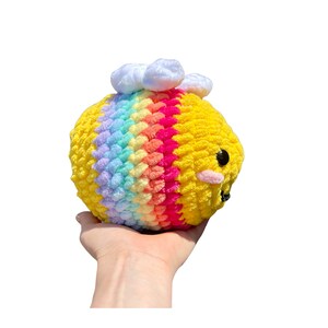 May include: A handmade, plush bee toy with a yellow body and a rainbow stripe across the middle. The bee has black eyes, pink cheeks, and white cloud accents on top. The toy is held in a hand against a white background.