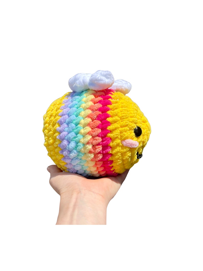 May include: A handmade, plush bee toy with a yellow body and a rainbow stripe across the middle. The bee has black eyes, pink cheeks, and white cloud accents on top. The toy is held in a hand against a white background.