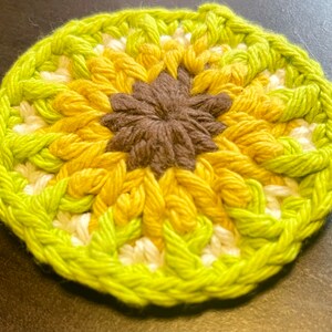 Pu&ograve; includere: Un sottobicchiere circolare all'uncinetto con un centro marrone scuro circondato da strati di petali in filato giallo e bianco, rifinito con un bordo esterno verde lime vibrante. Il pezzo tessile presenta un motivo strutturato ispirato ai fiori.