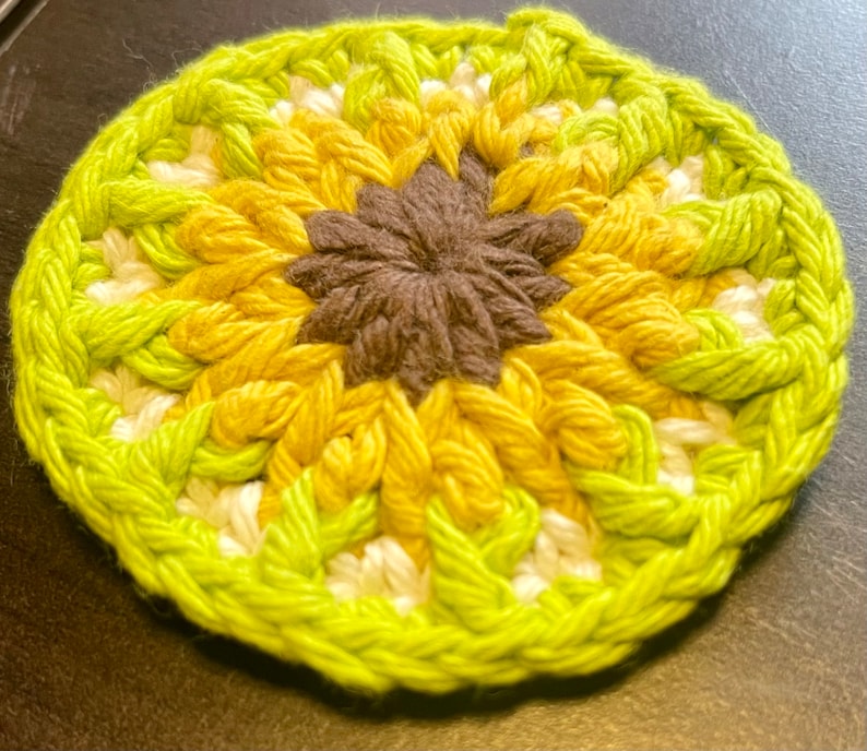 Pu&ograve; includere: Un sottobicchiere circolare all'uncinetto con un centro marrone scuro circondato da strati di petali in filato giallo e bianco, rifinito con un bordo esterno verde lime vibrante. Il pezzo tessile presenta un motivo strutturato ispirato ai fiori.