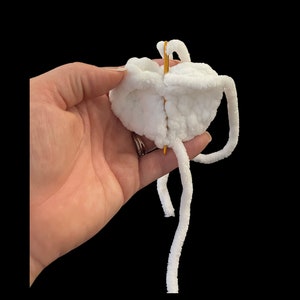 May include: A close-up of a white, fluffy, crocheted object held against a black background. A gold needle is inserted through the center of the object. The object has long, white, fuzzy strands extending from it.
