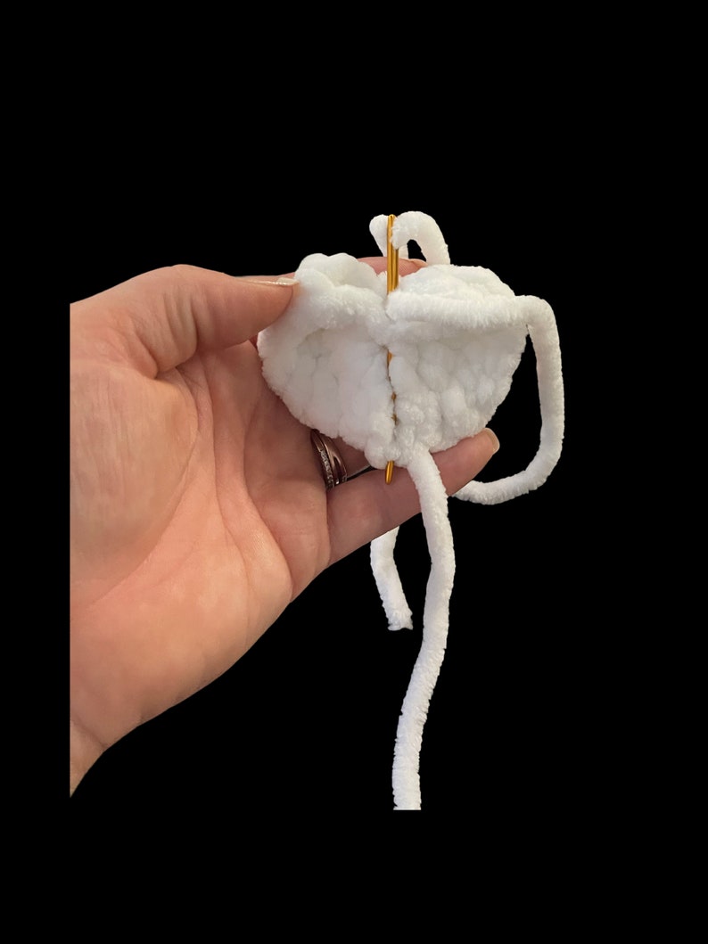 May include: A close-up of a white, fluffy, crocheted object held against a black background. A gold needle is inserted through the center of the object. The object has long, white, fuzzy strands extending from it.