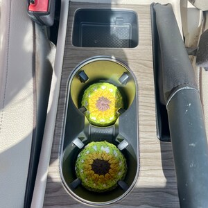 Pu&ograve; includere: Due sottobicchieri rotondi all'uncinetto con un design ispirato al girasole, caratterizzati da un centro marrone scuro circondato da strati di petali in filato giallo e verde chiaro, inseriti nei portabicchieri della console centrale.
