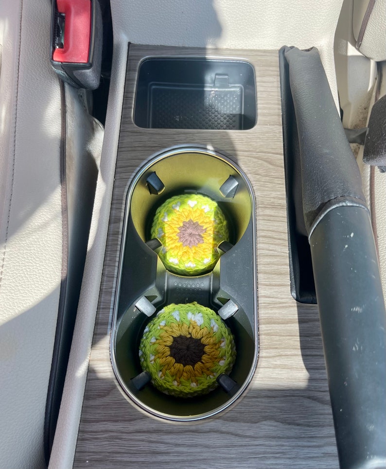 Pu&ograve; includere: Due sottobicchieri rotondi all'uncinetto con un design ispirato al girasole, caratterizzati da un centro marrone scuro circondato da strati di petali in filato giallo e verde chiaro, inseriti nei portabicchieri della console centrale.