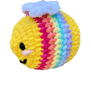 May include: A handmade, plush bee toy with a yellow body and rainbow-colored wings. The bee has black eyes, a pink mouth, and a white cloud on top. The toy is made of soft, textured yarn.