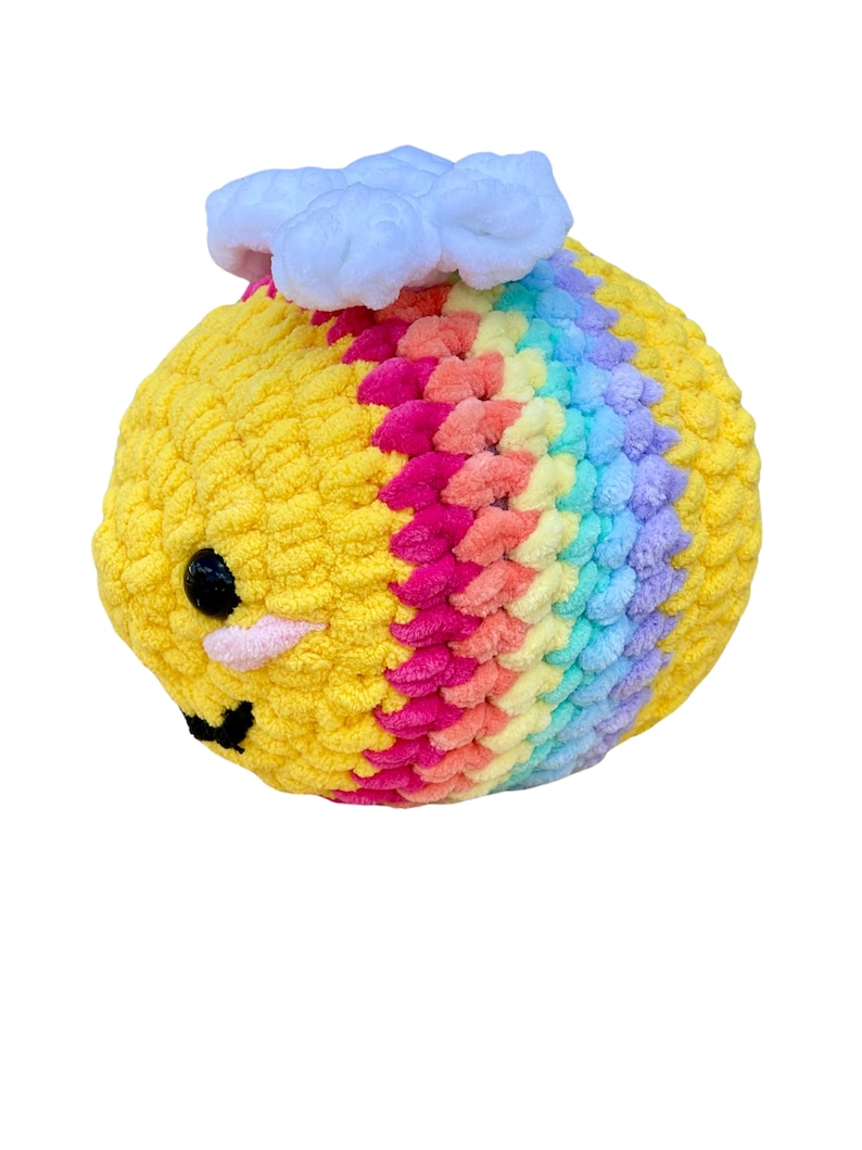 May include: A handmade, plush bee toy with a yellow body and rainbow-colored wings. The bee has black eyes, a pink mouth, and a white cloud on top. The toy is made of soft, textured yarn.