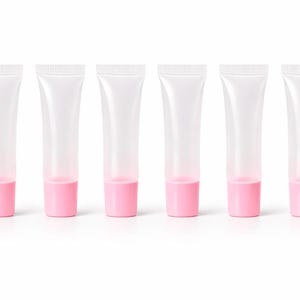 May include: Six clear, empty lip gloss tubes with pink bases are arranged in a row. The tubes are transparent, revealing the pink color at the bottom. The caps are white with a ridged texture.