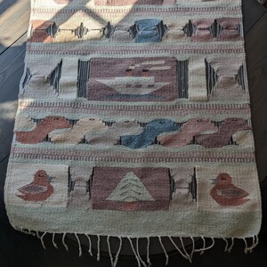 Pode incluir: Uma tape&ccedil;aria de parede tecida com um design geom&eacute;trico que apresenta p&aacute;ssaros, barcos e formas abstratas em tons de marrom, azul e branco. A tape&ccedil;aria tem uma borda de franjas.